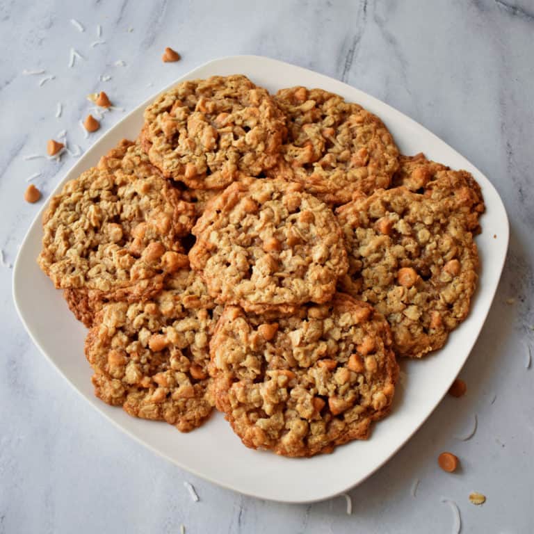 Overhead view of a square white plate filled with butterscotch oatmeal cookies with butterscotch chips and shredded coconut scattered around the plate.