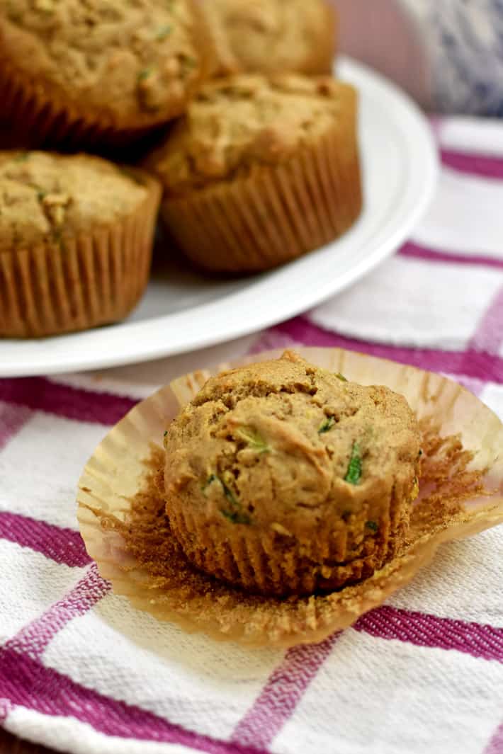 Close up of 1 gluten free zucchini muffin on a pink and white striped kitchen towel with more muffins on a white round plate in the background.
