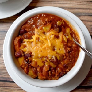 Overhead view of chili with beef, turkey and potatoes and a spoon in a white bowl sitting on a white plate.