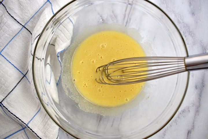 Overhead view of egg mixture for making gf zucchini bread in glass bowl with wire whisk and blue and white striped kitchen towel on the left.
