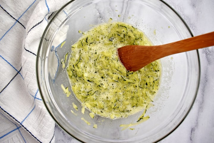 Overhead view of egg mixture, shredded zucchini, and butter for making gf zucchini bread in a glass bowl with a wooden spoon and a blue and white striped kitchen towel on the left.