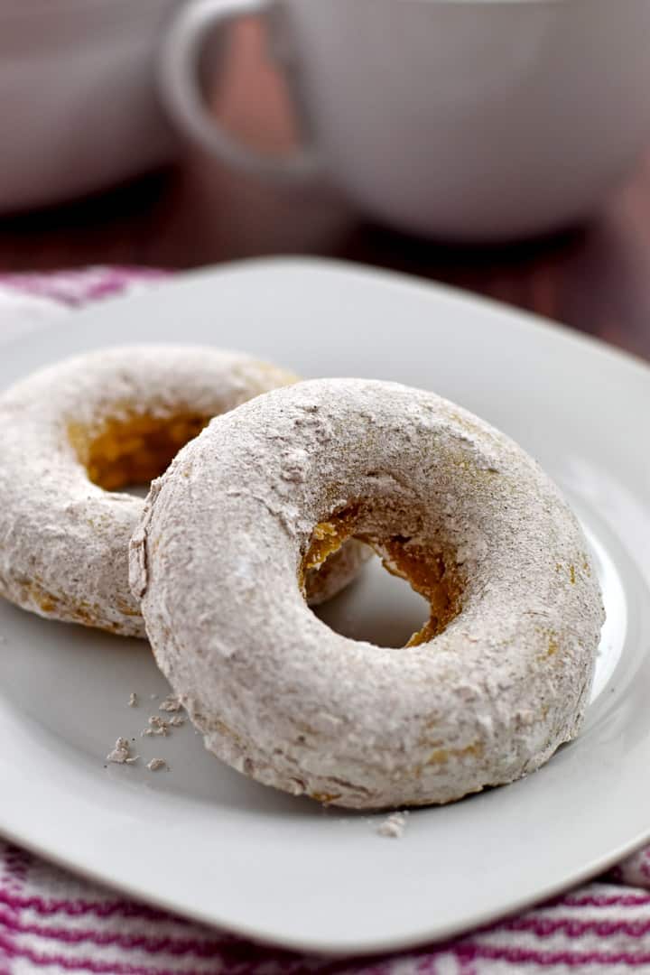 Two gluten free pumpkin donuts on a small white plate with a white coffee cup and bowl in the background.