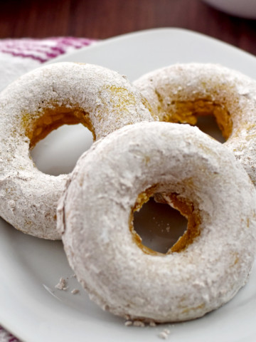 Three gluten free pumpkin donuts on a small white plate with a pink and white striped kitchen towel on the left.