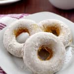 Three gluten free pumpkin donuts on a small white plate with a cup of coffee and a platter with more donuts in the background.