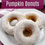 Three gluten free pumpkin donuts on white plate on top of a pink and white striped kitchen towel with a cup of coffee and platter of more donuts in the background. and a pink text overlay.