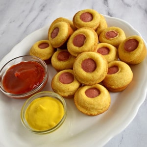 White oval platter filled with gluten free mini corn dog muffins with a side of mustard and ketchup in glass ramekins.