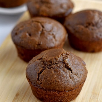 Four gluten free double chocolate muffins on a wood cutting board with more muffins in the background.