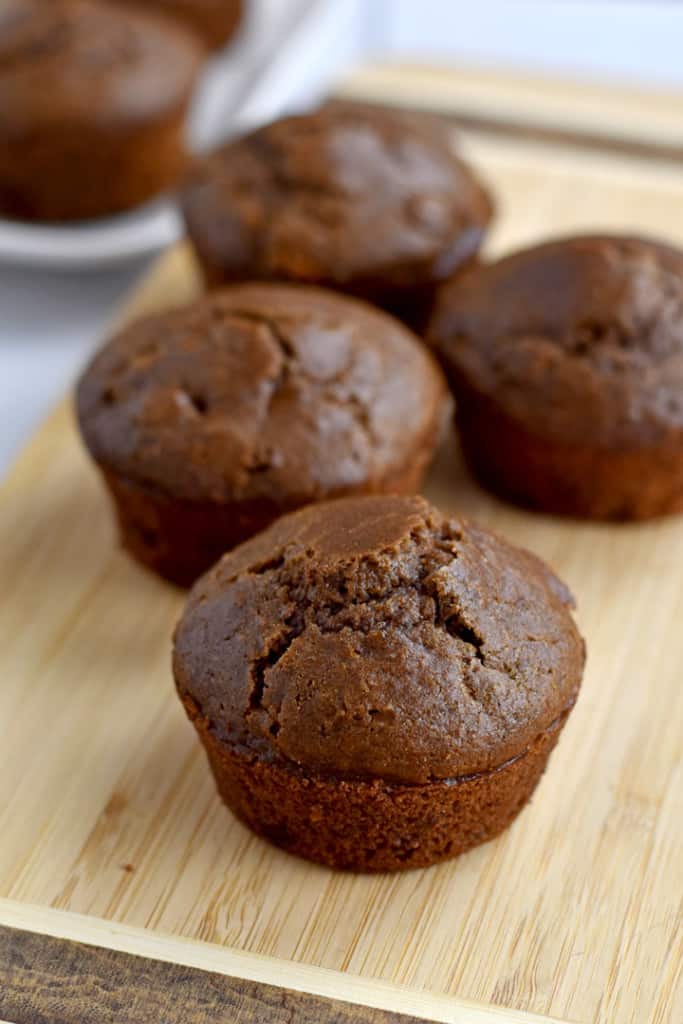 Four gluten free double chocolate muffins on a wood cutting board with more muffins in the background.