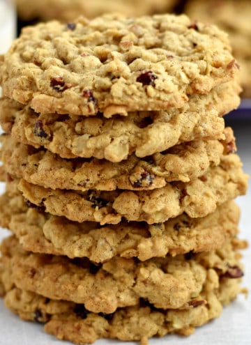 A stack of 7 gluten free cranberry oatmeal cookies on the counter with a glass of milk and a purple plate with more cookies in the background.