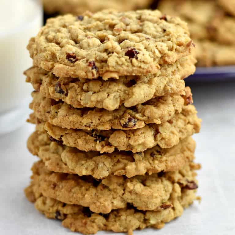 A stack of 7 gluten free cranberry oatmeal cookies on the counter with a glass of milk and a purple plate with more cookies in the background.