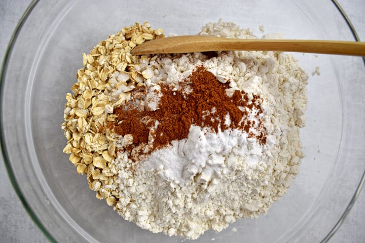Overhead view of glass mixing bowl with gluten free oats, gluten free flour, salt, baking soda, cinnamon, and a wooden spoon.
