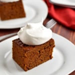 A slice of gluten free gingerbread cake, topped with whipped topping, on a small white plate with a fork and more cake in the background.