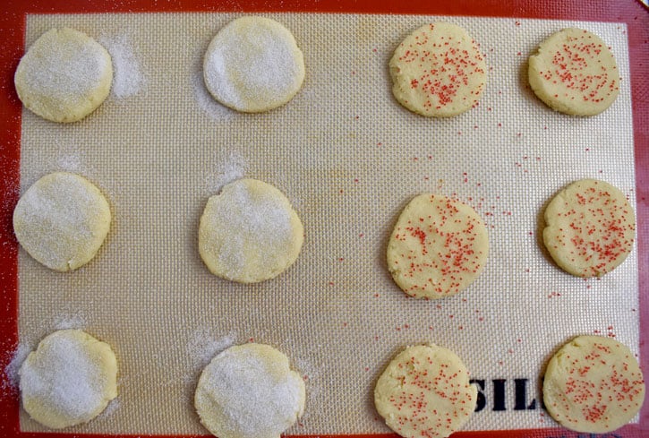 Overhead view of unbaked gluten free butter cookies on a silicone liner. The cookies on the left are sprinkled with sugar and the cookies on the right are decorated with red sprinkles.