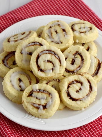 Overhead view of a round white plate, filled with gluten free cinnamon roll cookies, on a red and white checked kitchen towel.