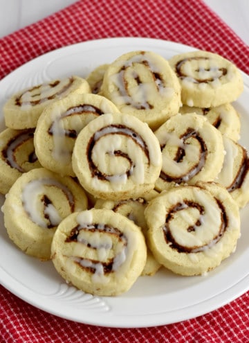 Overhead view of a round white plate, filled with gluten free cinnamon roll cookies, on a red and white checked kitchen towel.