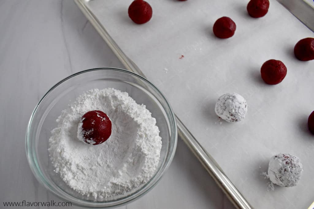 Overhead view of a small glass bowl with powdered sugar and a gluten free red velvet cookie dough ball waiting to be coated next to a parchment lined baking pan with more dough balls.
