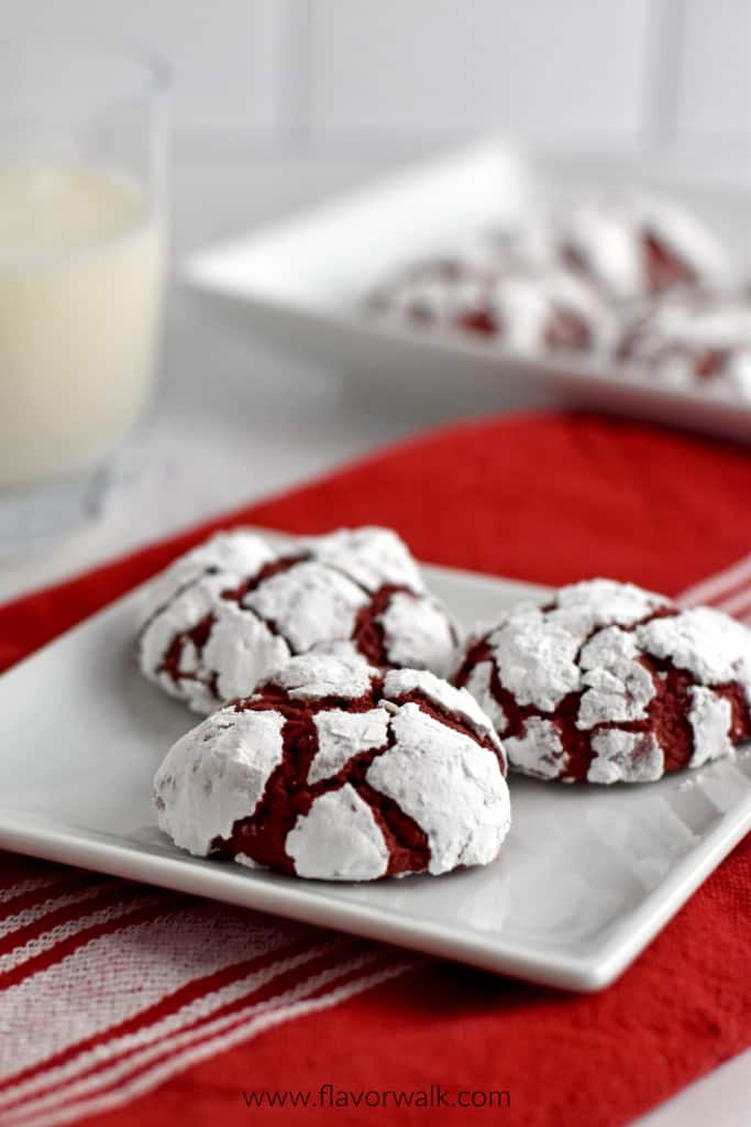 A square white plate with 3 gluten free red velvet crinkle cookies sitting on a red and white striped kitchen towel with a glass of milk and more cookies in the background.