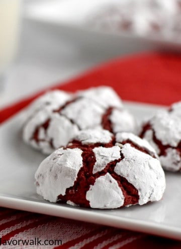 Three gluten free red velvet crinkle cookies on small white plate with red and white striped kitchen towel underneath plate. A glass of milk and more cookies in the background.