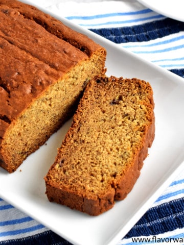 Overhead view of a loaf of gluten free peanut butter banana bread and one slice on a white rectangular serving platter sitting on a blue and white striped kitchen towel.