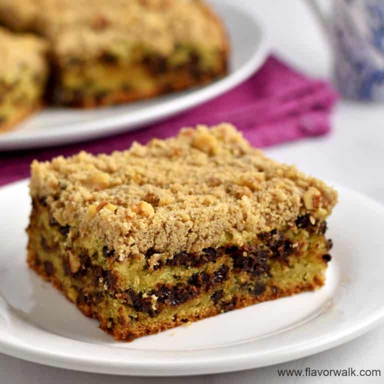 A piece of gluten free chocolate chip crumb cake on a small white plate with additional pieces of cake and a blue and white coffee cup in the background.