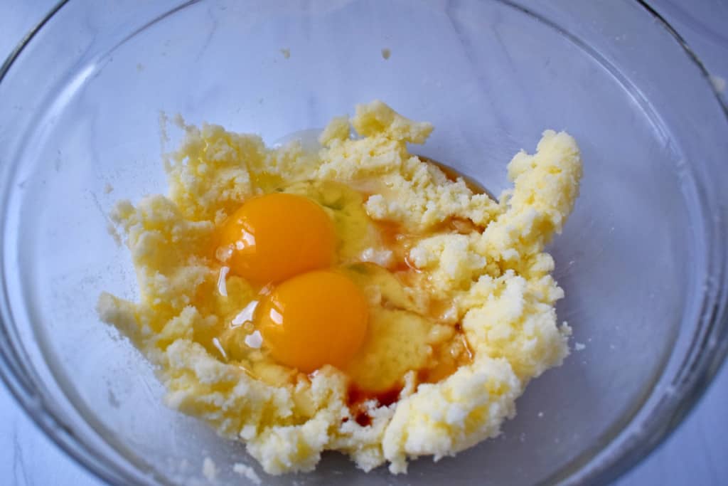 Overhead view of glass mixing bowl with butter, sugar, eggs, almond extract, and vanilla extract prior to being mixed together.