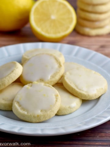 A small round plate filled with gluten free lemon cookies with lemons and stacks of more cookies in the background.