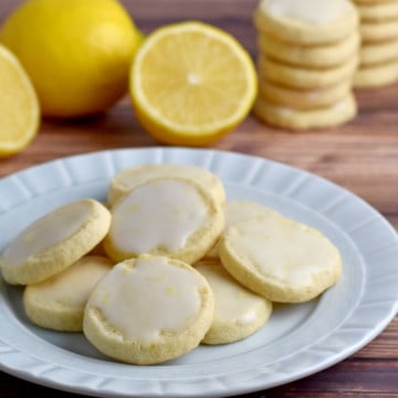 A small round plate filled with gluten free lemon cookies with lemon glaze sitting on a wooden table, with lemons and stacks of more cookies in the background.