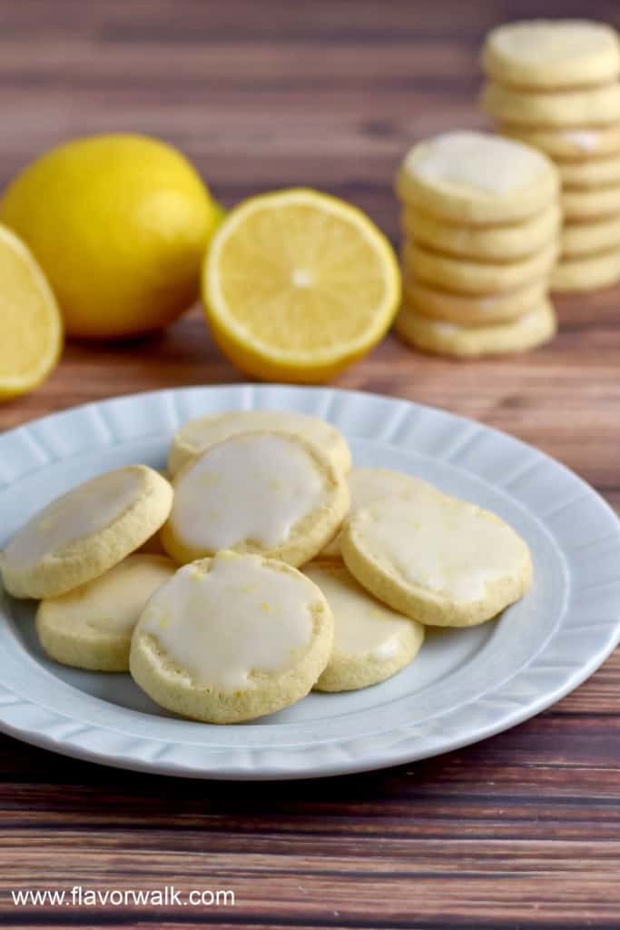 A small round plate filled with gluten free lemon cookies with lemon glaze sitting on a wooden table, with lemons and stacks of more cookies in the background.