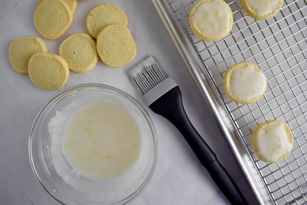 Overhead view of small glass bowl with lemon glaze and pastry brush with unglazed lemon cookies in the background and glazed lemon cookies on the right.