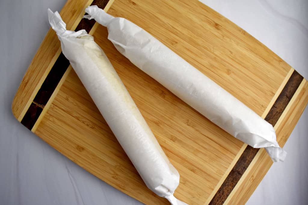 Overhead view of two rolls of lemon cookie dough rolled up in parchment paper sitting on a wooded cutting board.