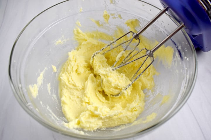 Large glass mixing bowl containing creamed together butter and sugar with electric hand mixer on the right.