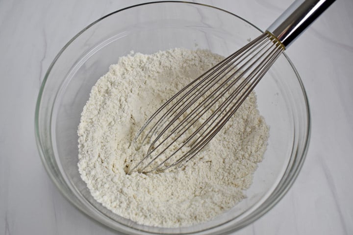 Overhead view of medium glass mixing bowl containing whisked together dry ingredients, gluten free flour blend, baking powder, and salt, and wire whisk.