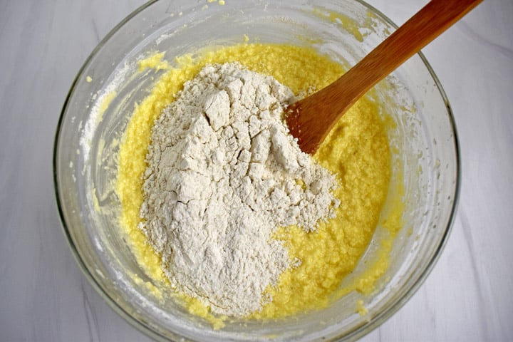Overhead view of glass mixing bowl containing ½ the dry ingredients for gluten free lemon pound cake being added to the wet ingredients with a wooden spoon.