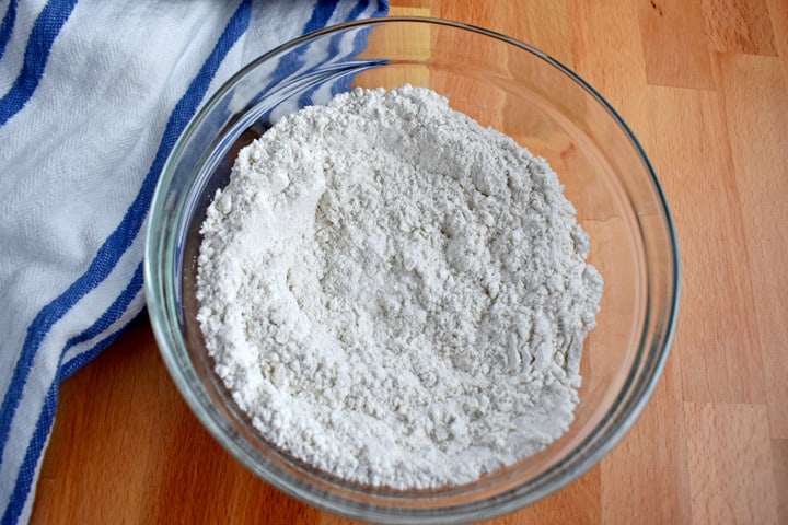 Overhead view of whisked together gf flour blend, baking powder, baking soda, and salt in medium glass mixing bowl with blue and white striped kitchen towel on the left.