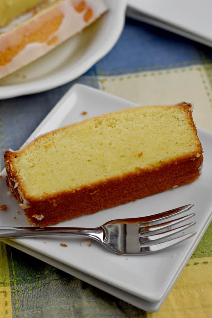 Close up view of a slice of gluten free lemon pound cake and a fork on a stack of 2 white dessert plates with more cake and plates in the background.
