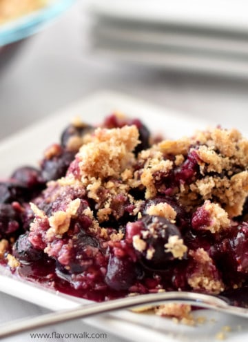 Close up view of a serving of gluten free blueberry crisp and a fork on a small white plate with more crisp and plates in the background.