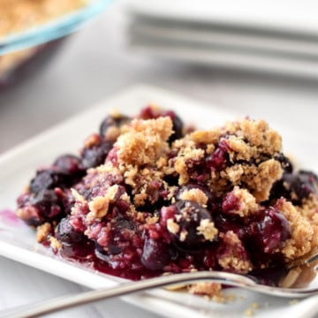 Small white plate filled with gluten free blueberry crisp and a fork with more blueberry crisp and plates in the background.