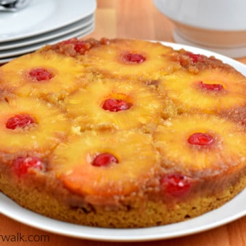 Close up view of pineapple upside-down cake on round white plate with dessert plates, forks, and coffee pot in background.