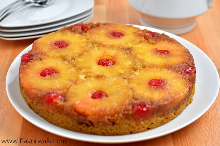 Close up view of pineapple upside-down cake on round white plate with dessert plates, forks, and coffee pot in background.