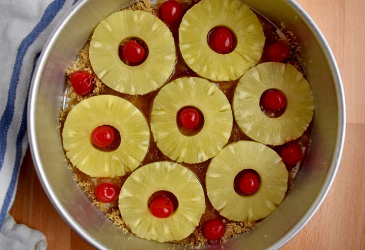 Overhead view of pineapple slices and maraschino cherries layered on top of butter and brown sugar in a 9x2-inch cake pan.