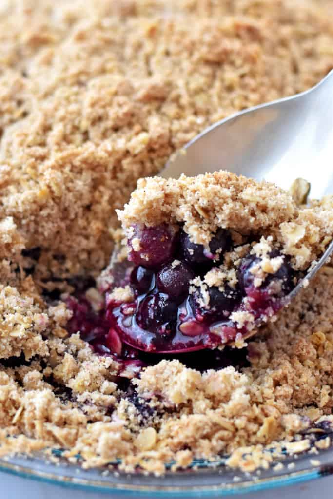 Close up view of a serving spoon lifting a scoop of gluten free blueberry crisp out of the pie dish.