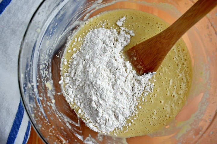 Overhead view of large glass mixing bowl with dry ingredients being stirred in to wet ingredients with a wooden spoon.