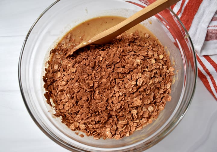 Overhead view of dry ingredients being added to wet ingredients for making chocolate peanut butter oatmeal cups and a wooden spoon in a glass mixing bowl.