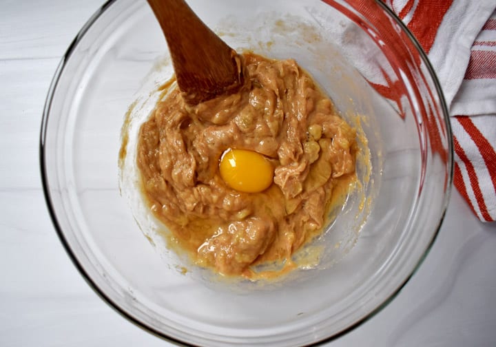 Overhead view of glass mixing bowl containing peanut butter/banana mixture an egg and a wooden spoon with a red and white striped kitchen towel on the right.
