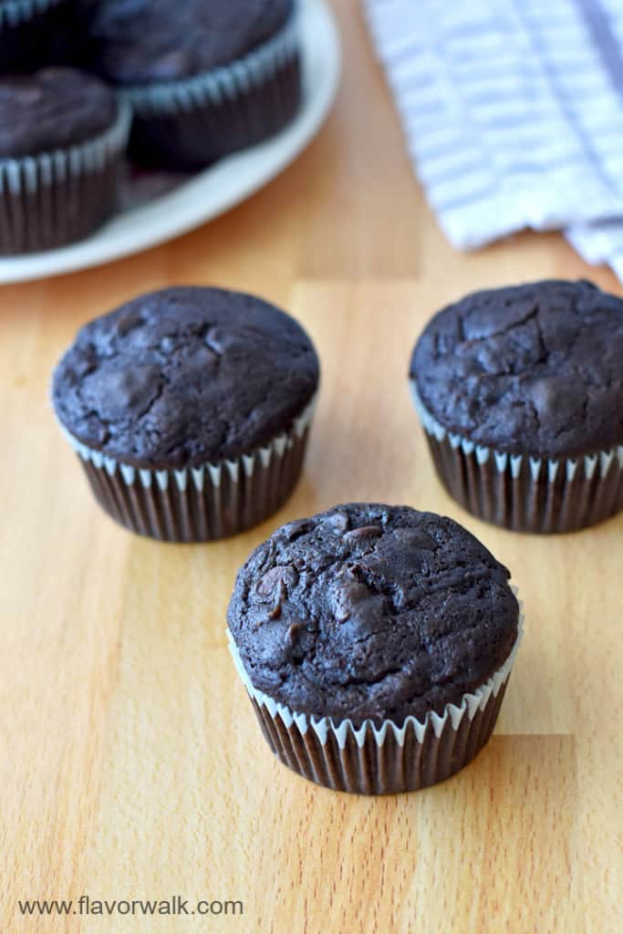 3 gluten free chocolate zucchini muffins on a wooden counter with more muffins and a purple and white striped kitchen towel in the background.