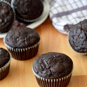 Gluten Free Chocolate Zucchini Muffins on a wood counter with more muffins on a plate and a purple and white striped kitchen towel in the background.