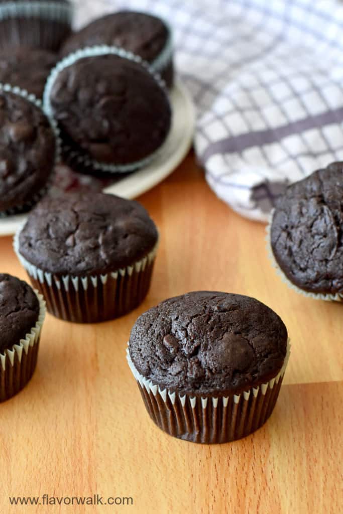 Gluten Free Chocolate Zucchini Muffins on a wood counter with more muffins on a plate and a purple and white striped kitchen towel in the background.