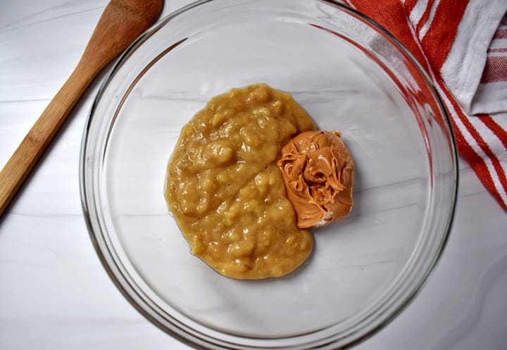 Overhead view of mashed bananas and creamy peanut butter in a glass mixing bowl with a wooden spoon on the left and a red and white striped kitchen towel on the right.
