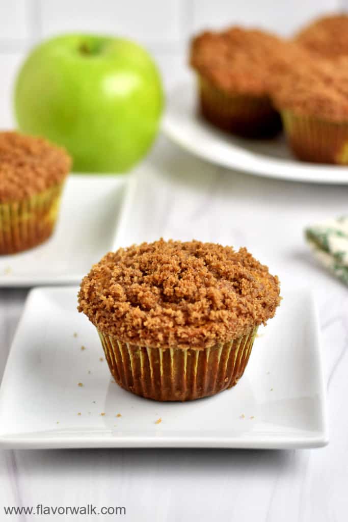 One gluten free apple muffin on a square white plate with more muffins and a granny smith apple in the background.