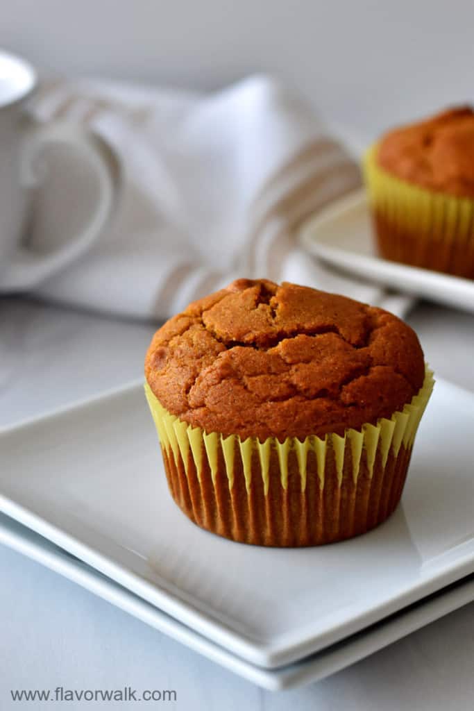 One gluten free pumpkin muffin on a stack of 2 small white plates with a white coffee cup, tan and white striped kitchen towel, and more muffins in the background.